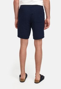 Granatowe szorty z teksturowanej tkaniny z tylną kieszenią, wykończone odwróconymi brzegami, noszone z ciemnymi sandałami.