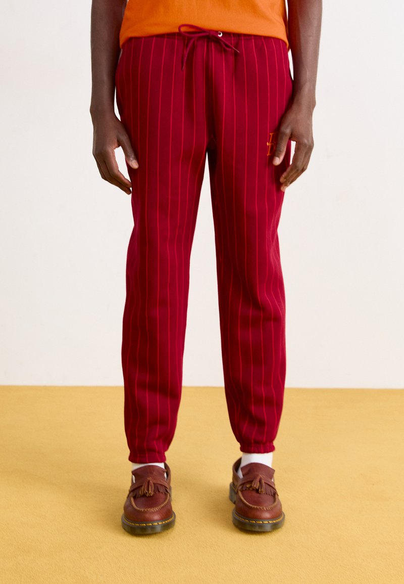 Red striped joggers with an elastic waistband and drawstring. Paired with brown slip-on shoes and white socks. Yellow background.