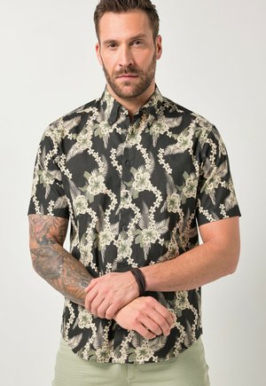 Uomo con capelli corti e barba che indossa una maglietta nera a maniche corte con motivi floreali e foglie beige, mostrando un braccio destro tatuato e mani giunte.