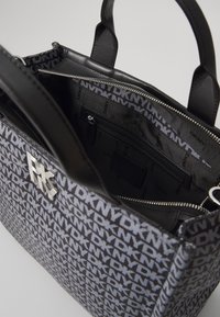 Bolsa de tecido preto com um padrão de logótipos repetidos, interior com fecho e um bolso, e duas alças. Inclui detalhe em metal prateado.