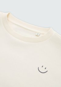 Felpa color crema con collo a girocollo a coste. Presenta una semplice grafica di un volto sorridente grigio sulla parte anteriore. Tessuto morbido.