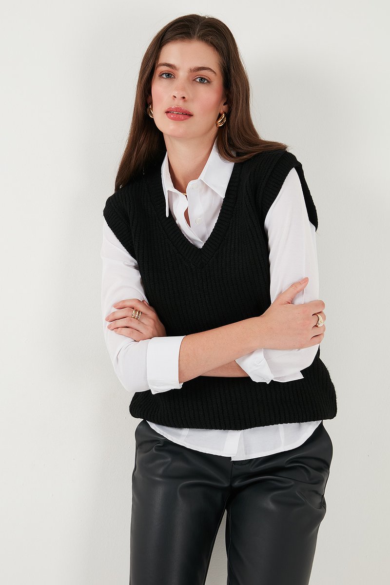 Sort strikket ærmeløs sweater med v-udskæring, båret over en hvid kravebluse. Modellen står med armene krydset og viser et struktureret mønster.