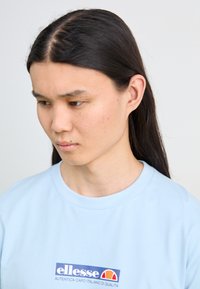 Light blue cotton t-shirt with a crew neck, featuring a logo patch in blue, white, and red at the chest. Smooth texture, casual fit.