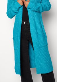 Cardigan lungo turchese con un motivo di maglia testurizzata, con polsini a coste e due tasche frontali. Indossato sopra pantaloni neri.