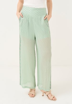 Femme portant un pantalon large vert clair texturé avec des panneaux latéraux à motifs et des sandales beiges à talons, les mains dans les poches.