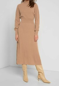 Beige stickad klänning med ribbat mönster, hög hals och långa ärmar. Matchas med beige spetsiga stövlar, vilket skapar en sammanhängande look.