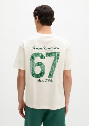Mann trägt ein weißes T-Shirt mit grüner Nummer „67“, dem Text „Scandinavian Football Community“ und „Marc O'Polo“ auf dem Rücken, kombiniert mit grünen Shorts.