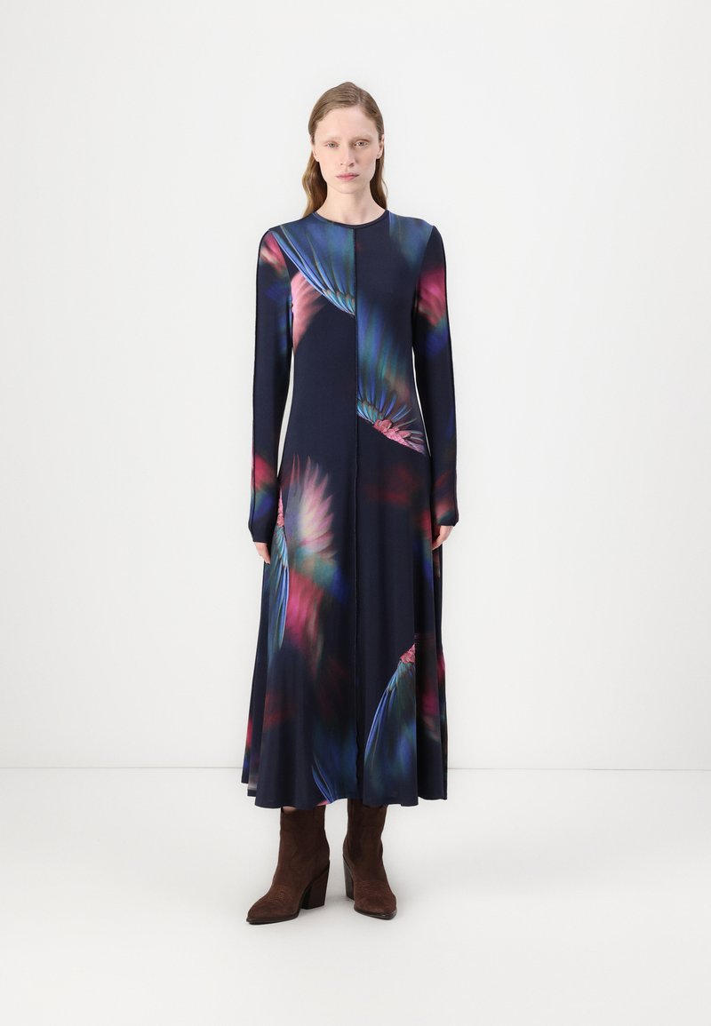 Rochie cu mâneci lungi de culoare bleumarin, cu un imprimeu floral vibrant în nuanțe de roz și verde, dintr-un material moale, având o siluetă fluidă. O pereche de cizme maro cu toc.