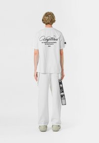 Camiseta blanca de gran tamaño con texto gráfico en la parte trasera en negro. Se combina con pantalones blancos holgados y calzado voluminoso de color claro.