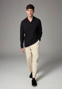 Svart långärmad tröja med V-hals, kombinerad med ljusa beige byxor och svarta slip-on skor. Modell står framför en grå bakgrund.
