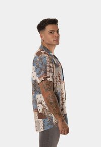 Camisa de manga corta en varios parches de azul, marrón y crema con patrones intrincados. Tejido suave y corte relajado, con cuello.