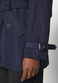 Giacca impermeabile blu navy con polsino con cintura, dotata di una grande tasca sulla spalla e chiusura con bottoni, realizzata in materiale liscio e lucido.