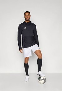 Juoda ilgomis rankovėmis sportinė viršutinė su aukštu apykakle, derinama su baltais šortais ir juodomis iki kelių kojinėmis, stovint ant juodos ir baltos futbolo kamuolio.