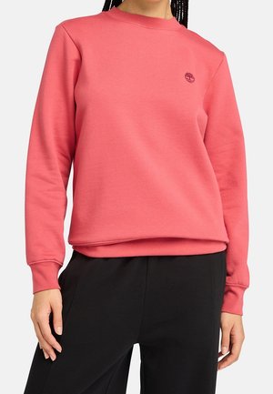 Kobieta w koralowo-różowej bluzie z długim rękawem z małym logo drzewa na lewej piersi i czarnych spodniach, stojąca na jednolitym tle.