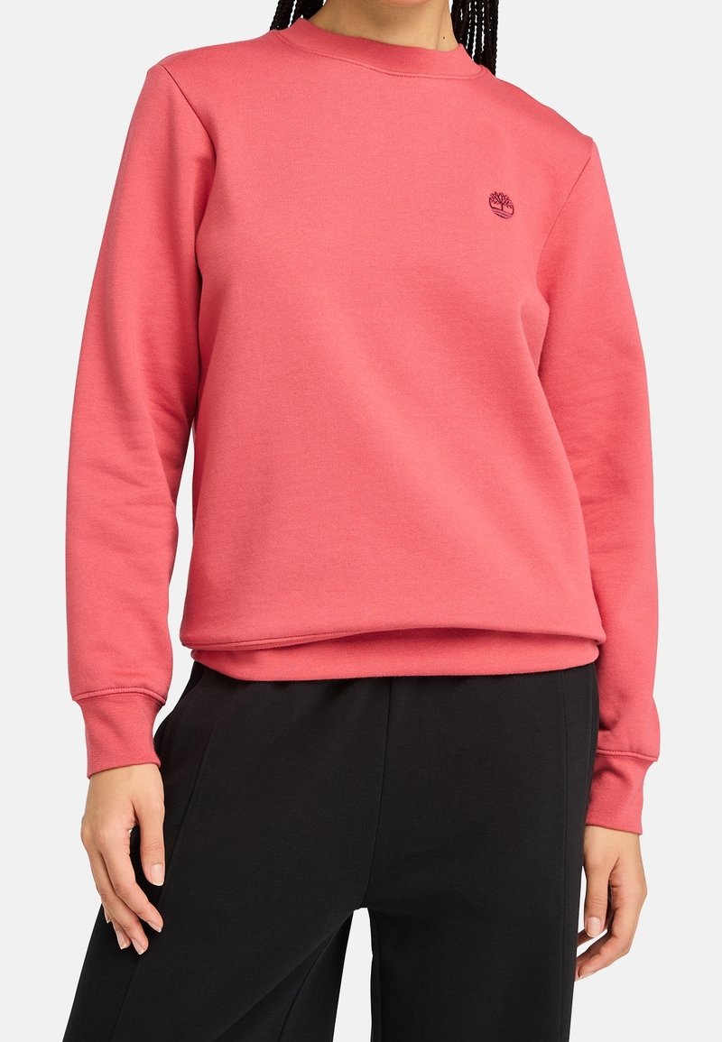 Femme portant un sweat-shirt corail rose à manches longues avec un petit logo d'arbre sur la poitrine gauche et un pantalon noir, debout devant un fond uni.