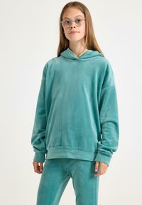 Turkusowa bluza z weluru z przednią kieszenią i lekko oversize'owym krojem, z kapturem na sznurki i ściągaczami na rękawach, w komplecie ze spodniami w tym samym kolorze.
