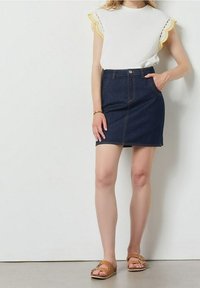 Džínová sukně s přiléhavým střihem, tmavě modré barvy a viditelnými švy. V kombinaci s bílou blůzkou s vykrojenými žlutými okraji.