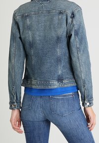 Giacca di jeans di un blu chiaro, con un taglio classico, polsini con bottone e dettaglio sulla parte posteriore. Indossata sopra un top blu e abbinata a jeans blu.