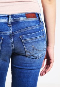 Jean en denim bleu avec une taille mi-haute, un design à cinq poches et une couture signature sur les poches arrière. Étiquette de marque en cuir tanné à la taille.