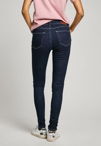 Sötétkék skinny farmernadrág magas derékkal, két hátsó zsebbel és kontrasztos sárga cérnával. Ezüst tornacipővel párosítva.