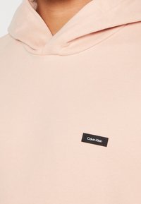 Felpa con cappuccio rosa chiaro in tessuto morbido, dotata di una piccola etichetta nera Calvin Klein sulla parte anteriore. Presenta un design del cappuccio rilassato.