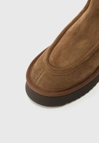 Brązowy zamszowy botek za kostkę z zaokrąglonym noskiem, wzmocnionym szwem i teksturowaną gumową podeszwą. Posiada subtelny wzór szwów.