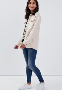 Beige Knopfjacke mit zwei Brusttaschen, getragen über einem schwarz-weiß gestreiften Oberteil, kombiniert mit blauen Skinny Jeans und weißen Sneakern.