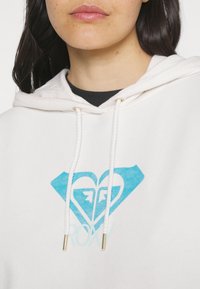 White hoodie featuring a blue graphic of a heart and diamond shape, with the text "ROXY" below in a lighter blue. Adjustable drawstrings with metallic tips.