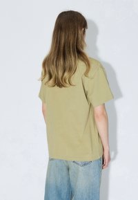 T-shirt di cotone verde oliva con maniche corte, vestibilità rilassata e scollo rotondo, abbinata a jeans di denim azzurro chiaro.