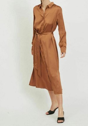 Kobieta nosząca satynową sukienkę w kolorze rdzy z długim rękawem, zapinaną na guziki, z paskiem w talii, w połączeniu z czarnymi, odkrytymi czubkami, damskimi butami na obcasie.