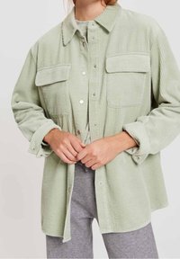 Donna che indossa una giacca a bottoni in corduroy verde chiaro con tasche a pattina sopra un top grigio e pantaloni grigi, con le mani unite all'altezza del ventre.