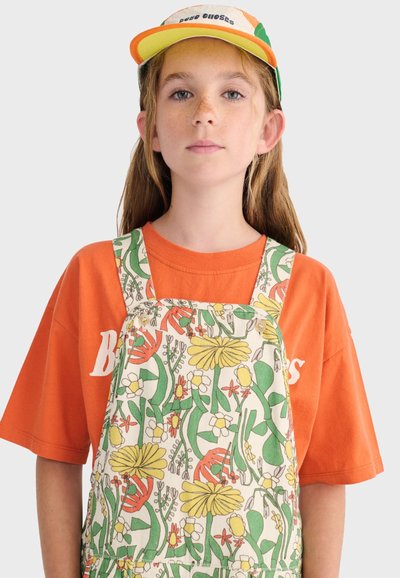 Jeune personne aux longs cheveux portant une chemise orange, une salopette à fleurs et une casquette multicolore sur un fond uni.
