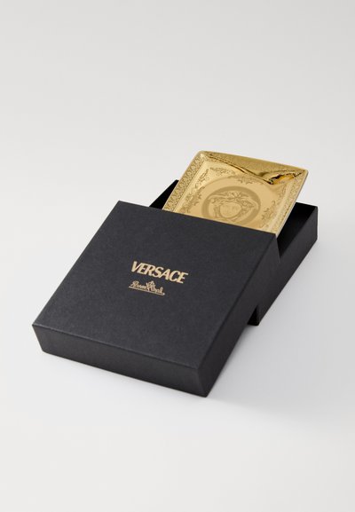 Χρυσό διακοσμητικό πιάτο με πολύπλοκες λεπτομέρειες, τοποθετημένο σε μαύρο κουτί με τη μάρκα Versace. Το πιάτο διαθέτει κεντρικό σχέδιο και περίτεχνες άκρες.