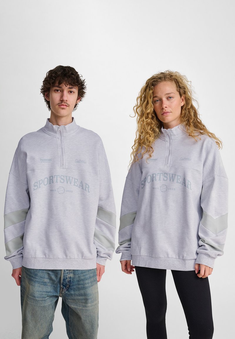 Grå zip-up sweatshirt med texten "SPORTSWEAR" tryckt i blått. Har långa, lösa ärmar och kontrasterande grå detaljer. Bärs över avslappnade byxor.