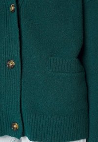 Tyrkysový vlněný cardigan s ribbovaným lemem a manžetami. Obsahuje zlaté knoflíky a náprsní kapsu vpředu, která ukazuje měkkou texturu.
