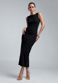 Black form-fitting sleeveless dress with a straight silhouette and a slight flare at the hem. Model wearing strappy gold sandals and accessories.