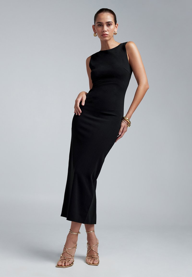 Black form-fitting sleeveless dress with a straight silhouette and a slight flare at the hem. Model wearing strappy gold sandals and accessories.