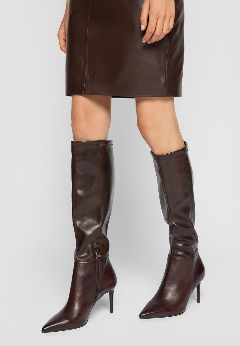 Botas de cuero marrón hasta la rodilla con puntera puntiaguda y tacón stiletto delgado; cuentan con un cierre de cremallera lateral y una textura suave.