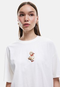 Camiseta de algodón blanca con un pato bordado que lleva una pajarita roja y zapatos, corte relajado con mangas cortas, textura suave y diseño casual.