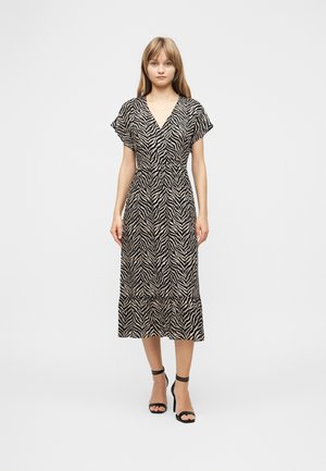 Kvinne iført en midikjole med svart og beige zebramønster, korte ermer og V-hals, kombinert med svarte hæler med ankelstropp, stående foran hvit bakgrunn.
