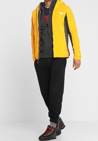 Yellow and gray insulated jacket with hood, layered over a dark gray sweatshirt. Black jogger pants and black boots with red accents.