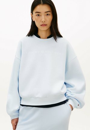 Ung kvinde med langt sort hår iført en lys blå oversize Tommy Hilfiger sweatshirt og matchende bukser mod en hvid baggrund.