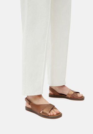 Brązowe skórzane sandały z fakturowanym paskiem, otwartym przodem i płaską podeszwą, w zestawieniu z białymi spodniami o szerokich nogawkach.