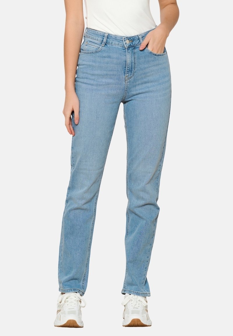 Femme portant un jean bleu clair droit avec des baskets blanches, debout avec une main dans la poche et l'autre détendue sur le côté.