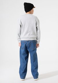 Sudadera gris de manga larga, cuello redondo y puños de canalé, combinada con unos vaqueros azules de corte holgado. El modelo lleva un gorro de punto negro.