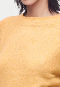 Primo piano di una persona che indossa un maglione morbido e arancione chiaro a maglia con scollatura rotonda, che mostra parte del collo e della spalla.