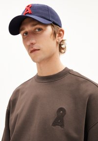 Sweat-shirt marron avec un accent logo texturé sur la poitrine, associé à une casquette bleu marine avec une lettre rouge brodée. Vue rapprochée du visage.