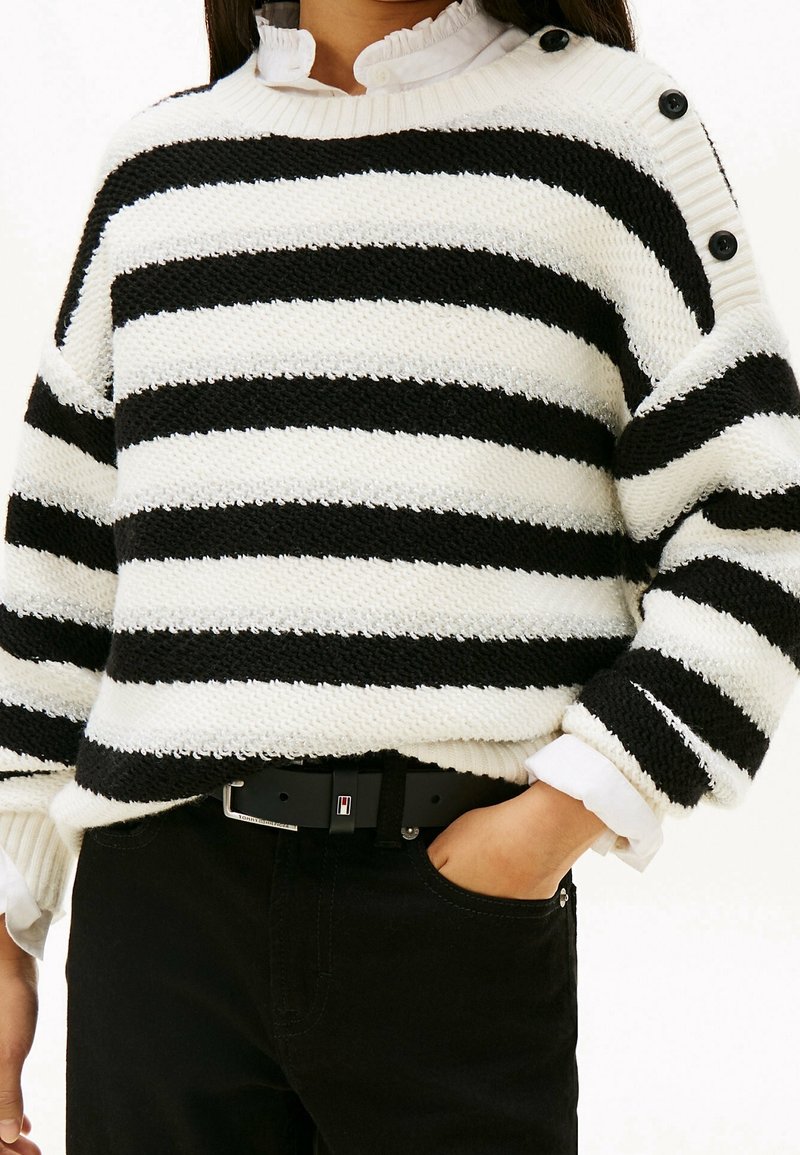 Femme portant un pull rayé noir et blanc avec des boutons à l'épaule, une chemise blanche en dessous, un pantalon noir, et une main dans la poche.