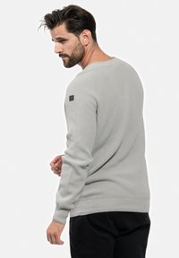 Pull en maille gris à col rond et manches longues ; il présente une petite étiquette noire sur l'épaule gauche et un ourlet ajusté.