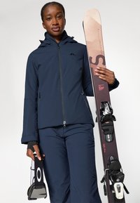 Chaqueta de esquí azul marino con cremallera en la parte delantera y capucha, combinada con pantalones a juego. Sosteniendo un par de esquís con fijaciones negras y un palo de esquí.